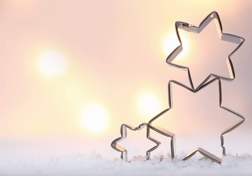 Atmospheric Christmas star background with three metal star cookie cutters balanced on top of each other on snow against a soft moody bokeh of glowing lights with copyspace