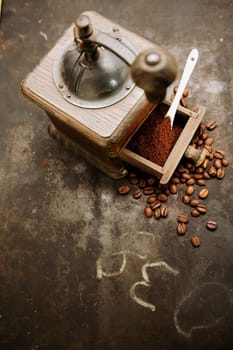 Overhead view of an old manual rustic wooden coffee grinder with an open drawer filled with freshly ground coffee with scattered roasted coffee beans on a grungy slate with scribblings and copyspace