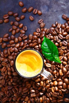 Overhead view of a cup of rich frothy coffee standing in scattered whole roasted coffee beans with a green leaf for decoration