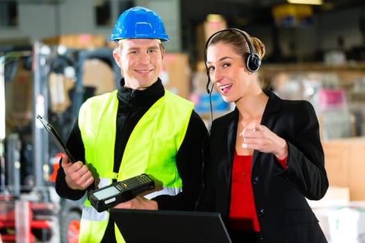 Teamwork - warehouse clerk or warehouse worker and customer service representative using laptop and headset in a warehouse of a freight forwarding company,she points at the camera