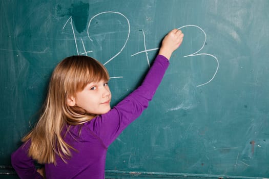 Young girl writing numbers on chalkboard with chalk while looking towards class