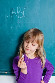 Young girl standing in front of chalkboard with letters of alphabet and looking at hand holding stick of chalk