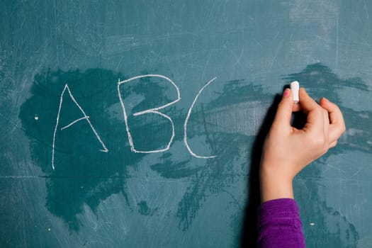 Hand of young child holding stick of white chalk to write letters of alphabet on chalkboard