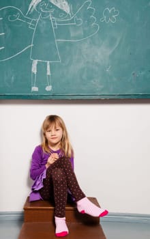 Studio portrait of relaxed young girl in classroom sitting in front of chalkboard with drawing of angel