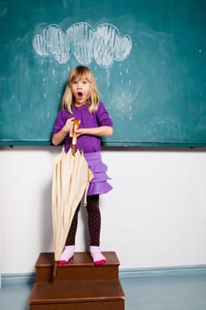 Studio portrait of surprised looking young girl holding an umbrella while standing in front of chalkboard with image of rain cloud