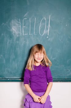 Studio portrait of smiling young girl with eyes closed in classroom with word hello written on chalkboard