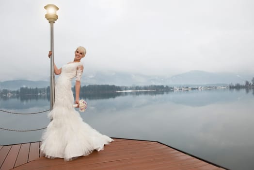 Young blonde bride in long white dress standing at a lantern at Lake Wolfgang