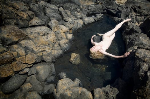 Nude of a blonde woman with fair skin in a pool in lava rock