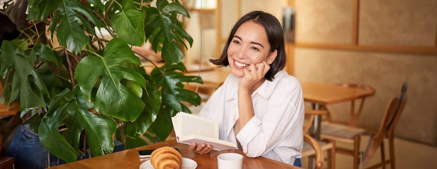 Romantic asian woman sitting with book in cafe, eating croissant and drinking coffee, reading and smiling, enjoying alone time.