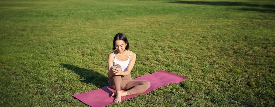 Woman in park, watching yoga video on smartphone, meditating on fresh air, sitting on rubber mat.