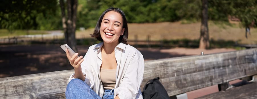 Portrait of happy young asian woman talking on mobile phone in park, sitting on bench and having a telephone call, chatting lively.