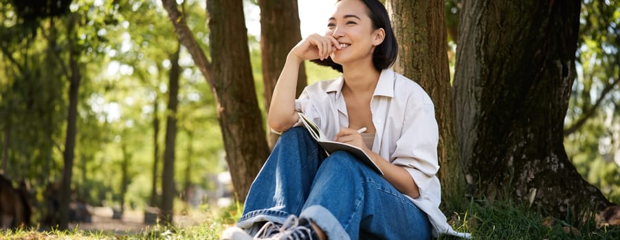 Portrait of young asian woman writing in her notebook, expressing her thoughts on paper in diary, smiling and sitting under tree on summer day in park.