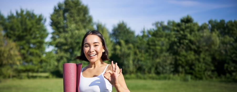 Excited young woman standing with sports mat, yoga clothes, shows okay sign, workout in park, wellbeing training session outdoors.