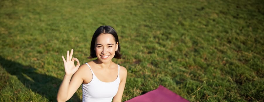 Portrait of asian woman sitting on rubber mat, meditating, showing okay sign and looking at camera, doing yoga.