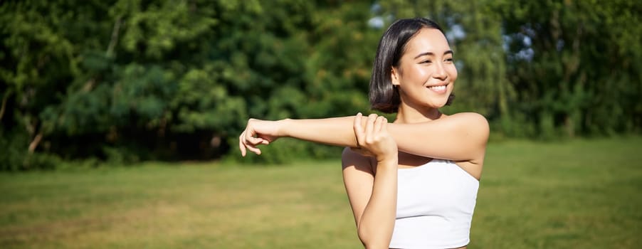 Portrait of young fitness woman stretching her arms, warm-up before training session, sport event in park, jogging and excercising.