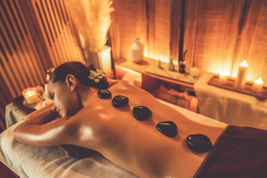 Hot stone massage at spa salon in luxury resort with warm candle light, blissful woman customer enjoying spa basalt stone massage glide over body with soothing warmth. Quiescent