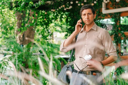 Young handsome man walk out of coffee shop with cafe garden, holding a coffee cup and a shopping bag while talking on the phone. Modern happy carefree with sunglasses lifestyle. Expedient