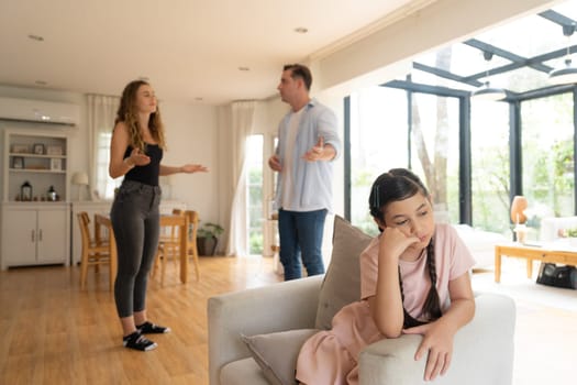 Annoyed and unhappy young girl sitting on sofa trapped in middle of tension by her parent argument in living room. Unhealthy domestic lifestyle and traumatic childhood develop to depression.Synchronos