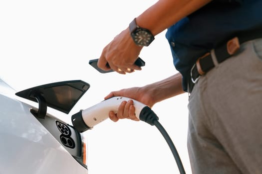 Young man use smartphone to pay for electricity at public EV car charging station at city commercial mall parking lot. Modern environmental and sustainable urban lifestyle with EV vehicle. Expedient