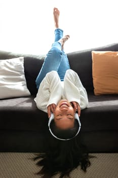 Happy young Chinese woman relaxing at home, lying on the couch upside down, listening to music with headphones. Vertical image. Lifestyle concept.
