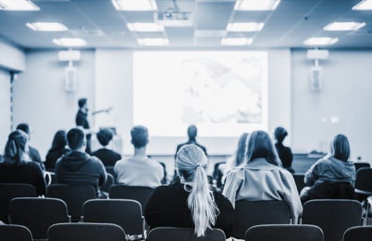 Speaker giving a talk in conference hall at business event. Rear view of unrecognizable people in audience at the conference hall. Business and entrepreneurship concept. Blue tones black and white