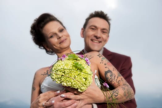 A bride with tattoos shows her bridal bouquet,behind it her groom