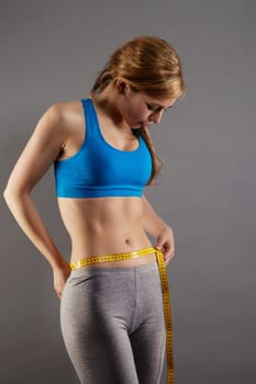 Pretty sportswoman measuring her waist against gray background