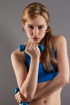 Young serious sportswoman with fitness dumbbells against gray background