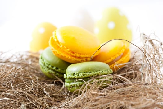 Close-up of colorful macaroons resting in straw nest and Easter eggs on light background