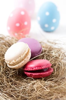 Close-up of colorful macaroons resting in straw nest and Easter eggs on light background