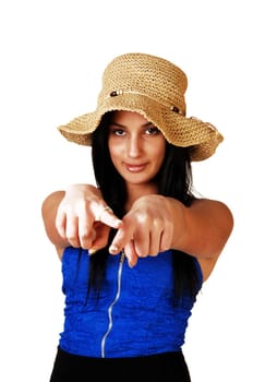 A teenager girl in a blue corset, black skirt and a straw hat standing for white background with her long black hair, pointing with both fingers at the photographer.