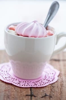 Closeup of cup of hot chocolate with pink meringue and spoon resting on small pink table mat on wooden table