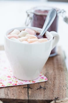Closeup of cup of hot chocolate with coloured marshmallows resting on serviette on wooden table with glass jar of cocoa powder in background