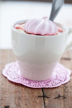 Closeup of cup of hot chocolate with pink meringue and spoon resting on small pink table mat on wooden table