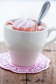 Closeup of cup of hot chocolate with pink meringue and spoon resting on small pink table mat on wooden table