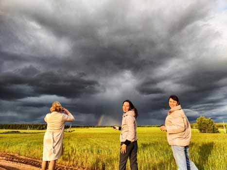 Tourist girls take pictures of rainbow and dramatic, gloomy clouds over a field on a spring, summer, autumn day. The concept of the beauty of nature and travel