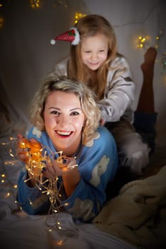 Cute mother and daughter in pajamas lie on soft blanket and having fun in room with Christmas garlands and white background. Tradition of decorating house for holiday. Happy childhood and motherhood