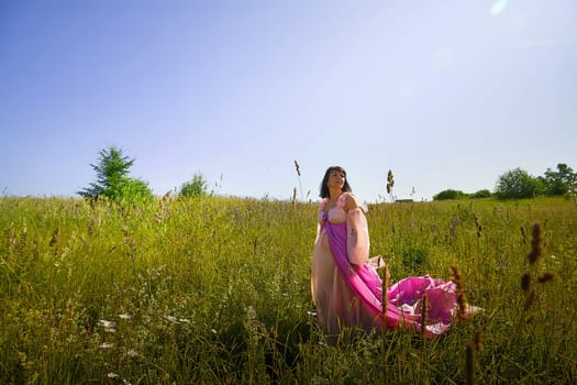 Beautiful girl in lush pink ball gown in green field during blooming of flowers and blue sky on background. Model posing on nature landscape as princess from fary tale