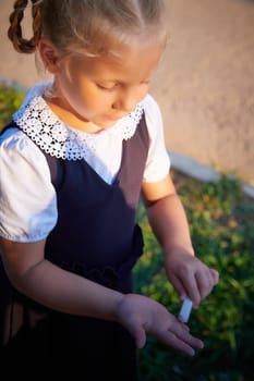 Little girl of elementary school student in modern school uniform drawing with chalk on asphalt outdoors. Female child schoolgirl going to school. Back to school in september 1