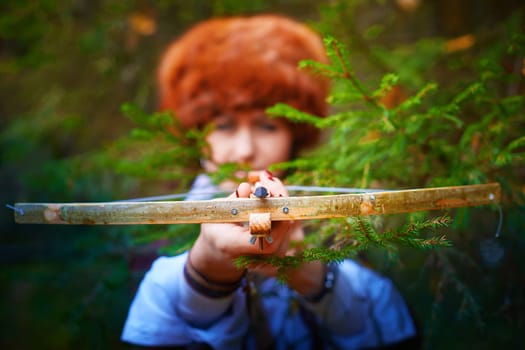 Girl in a leather jacket, a big red fox fur hat and with a crossbow in the forest in autumn. A female model poses as a fabulous royal huntress on nature hunt at a photo shoot