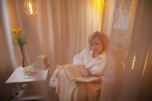 Adult mature woman of 40 or 60 years old with book in casual dress white shirt and blue jeans in calm cozy evening atmosphere room. Interior with transparent curtains and soft warm lamp