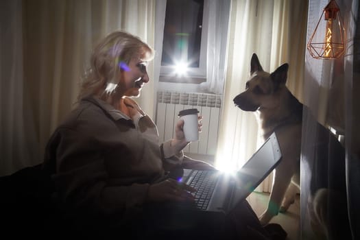 Adult mature woman of 40-60 with big shepherd dog in warm sweater. Room with calm cozy evening atmosphere with transparent curtains and soft warm light of lamps. Concept of love for animals and pet