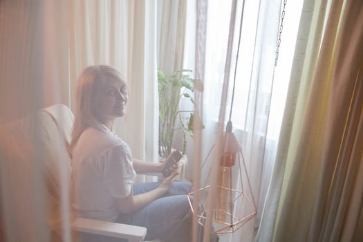 Young blonde woman using cell phone at home. Girl is resting, chating and looking in camera on smartphone near window at home. Nice atmosphere. The concept of cozy home and communication by Internet