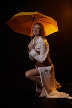 Beautiful sexy skinny girl in a dark hall with splashes of water and yellow umbrella under rain. Female Model blonde woman posing with water on black background in the studio