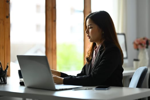 Asian woman economist in black suit working on laptop in bright office.