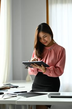 Charming asian businesswoman standing at her workplace and using digital tablet.