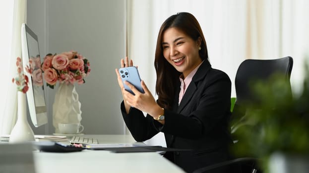 Cheerful millennial woman office worker waving hand, making video calling on smartphone.
