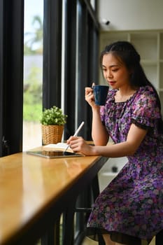 Young asian woman dressed casually drinking coffee and working on digital tablet at coffee shop.
