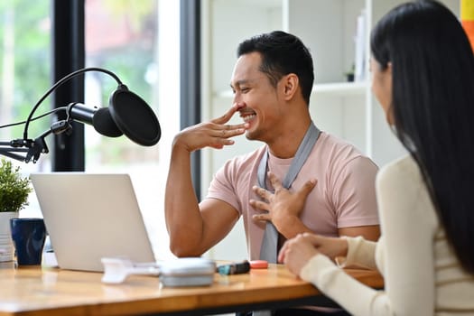 Cheerful gay male radio host discussing various topics with guest while streaming live audio podcast from studio.