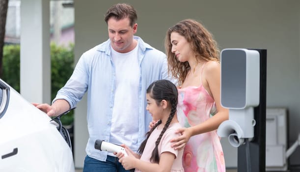 Happy little young girl learn about eco-friendly and energy sustainability as she help her family recharge electric vehicle from home EV charging station. EV car and modern family concept. Synchronos